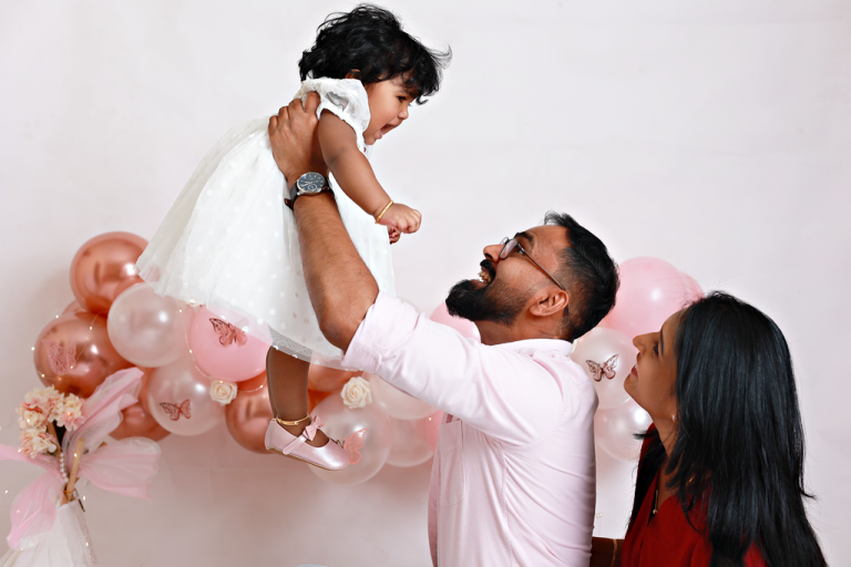 Father lifting his baby daughter into the air while mother smiles, pink balloon backdrop