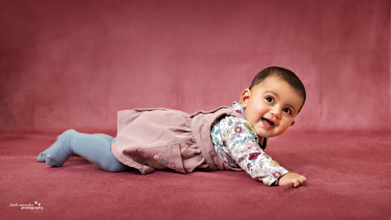Smiling baby girl lying on her tummy getting ready to crawl on a pink backdrop, baby milestone photoshoot in Doha