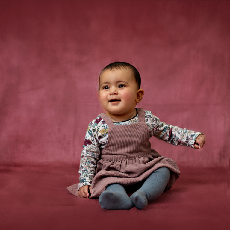 Smiling baby girl sitting in a pinafore dress on a pink backdrop, sitter session in Doha