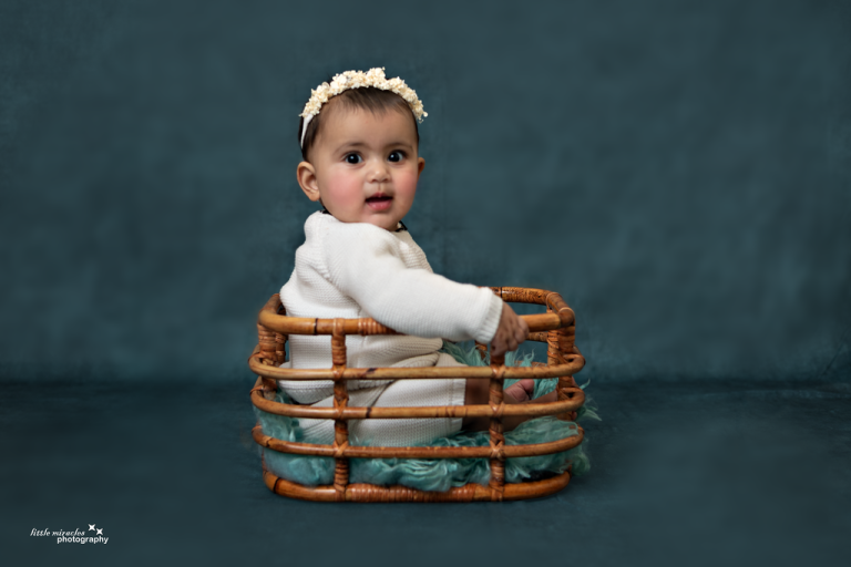 Baby with a flower headband sitting in a woven basket on a teal backdrop