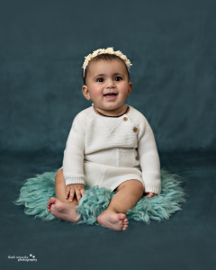 Smiling baby sitting on a teal fur rug in a cream knit outfit