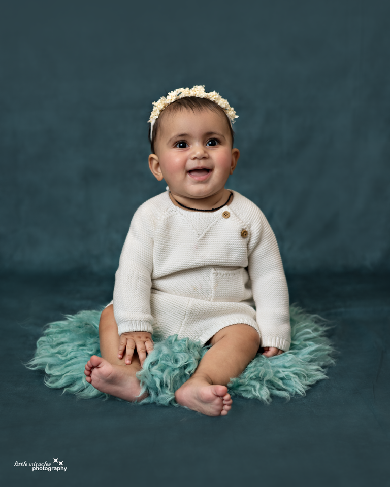 Smiling baby sitting on a teal fur rug in a cream knit outfit