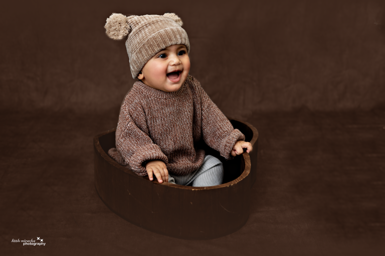 Laughing baby in a knitted hat sitting in a brown wooden bowl