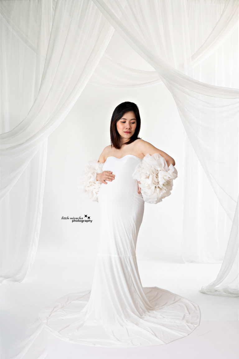 Mum-to-be in a white maternity gown with draped white curtains behind her