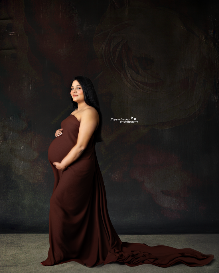 Pregnant woman in a fitted brown gown with a long train on a dark backdrop