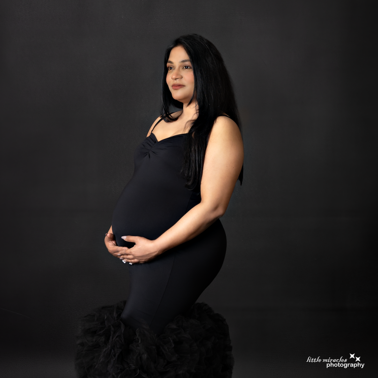Pregnant woman in a fitted black maternity dress holding her bump against a dark backdrop, maternity photoshoot in Doha