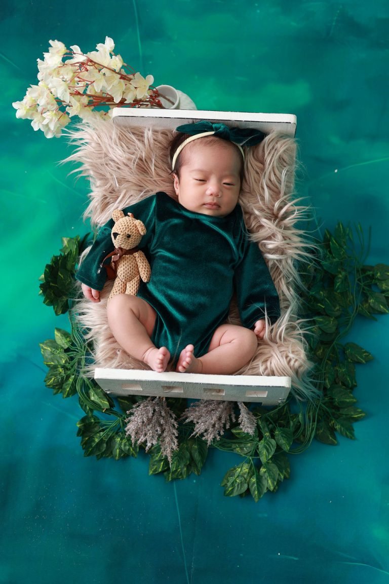 Newborn baby in a green outfit asleep in a white wooden crib with a teddy bear, newborn photography in Doha