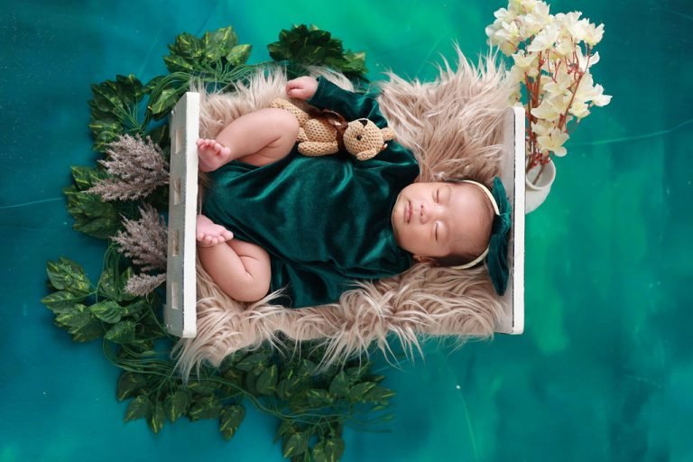 Newborn baby sleeping in a white crib with pampas grass and a teddy bear on a green backdrop