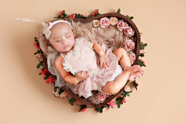 Sleeping newborn girl in a pink dress inside a heart-shaped bowl decorated with roses