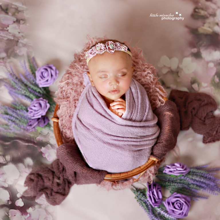 Newborn baby wrapped in lilac with a flower headband, surrounded by purple roses and lavender
