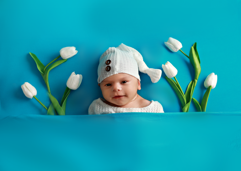 Baby in a knitted hat peeking out between white tulips on a blue backdrop