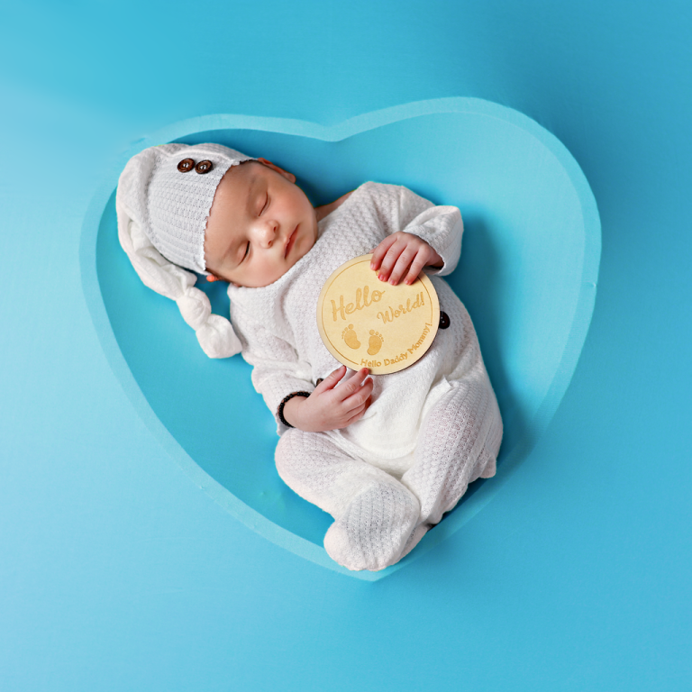 Newborn baby in a white outfit holding a "hello world" sign in a blue heart-shaped bowl