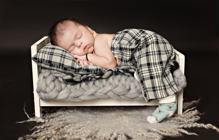 Newborn baby asleep on a small white bed with a checked blanket on a dark backdrop