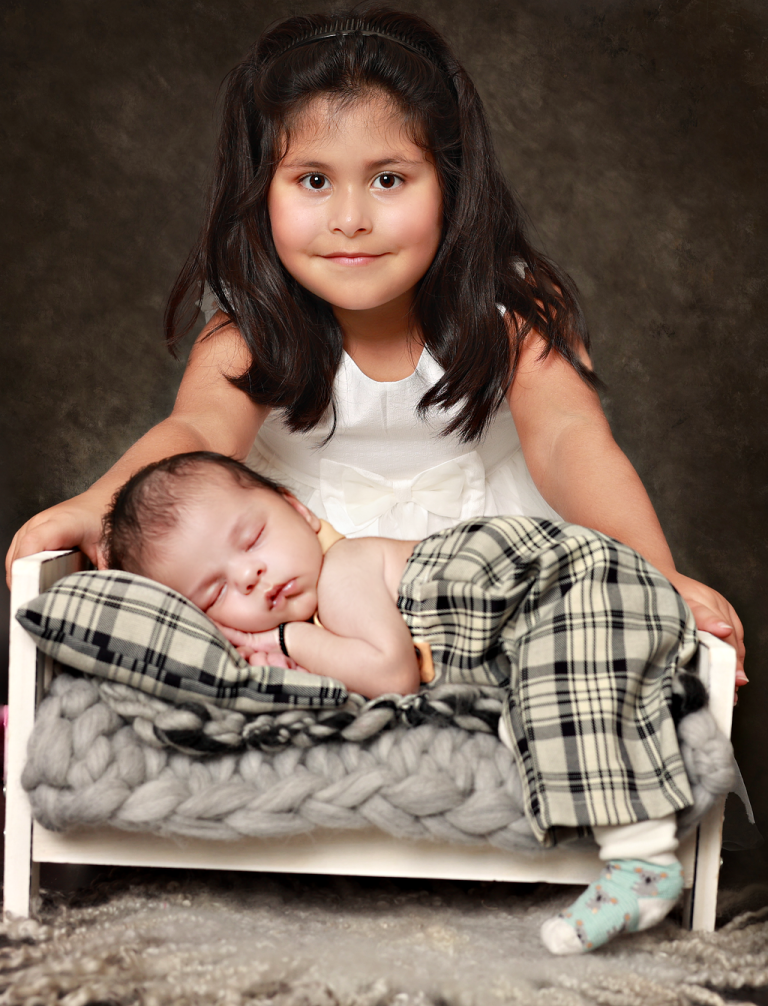 Big sister smiling behind her newborn sibling sleeping on a small bed
