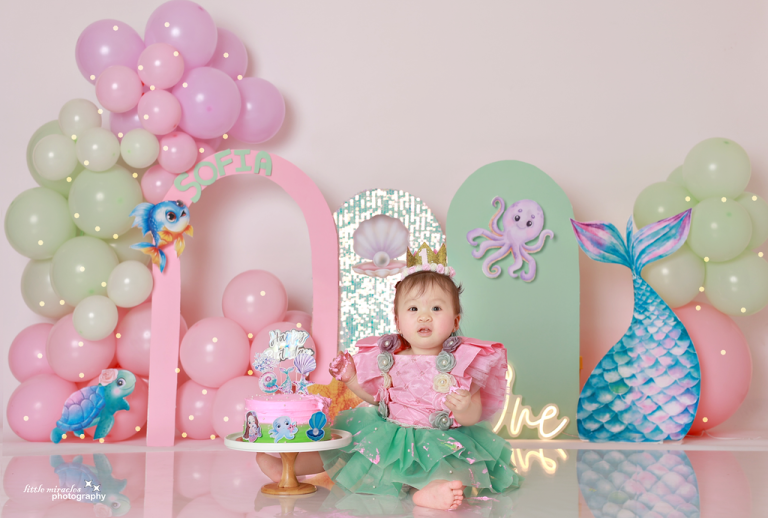 Baby girl in front of a mermaid-themed first birthday setup with pink and green balloons