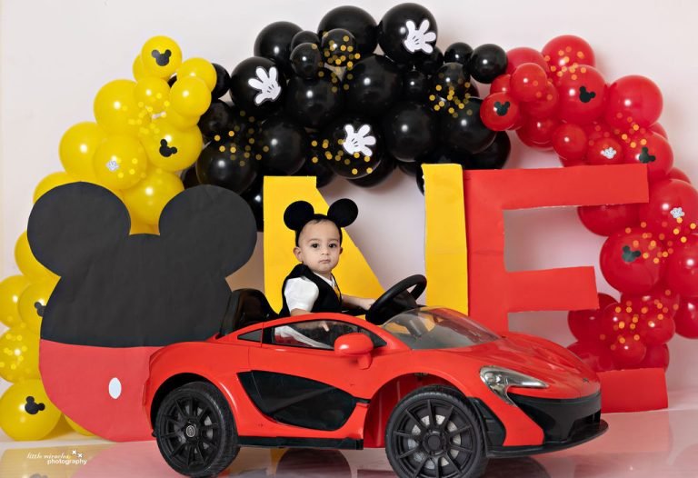 Baby boy in a red toy car at a Mickey Mouse first birthday setup