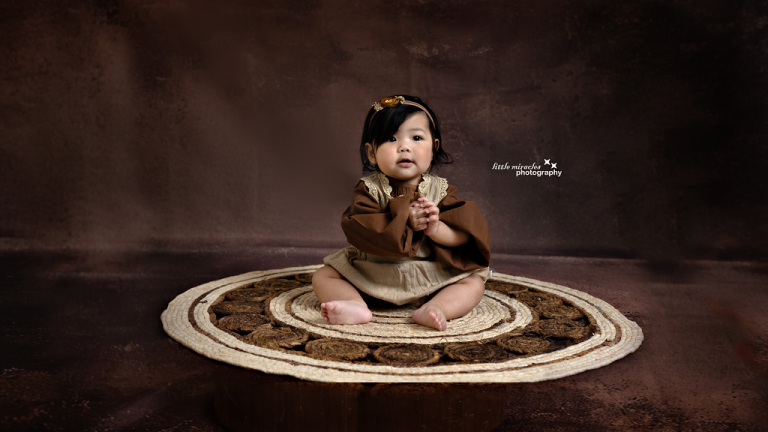 Baby girl sitting on a round rug against a dark brown backdrop