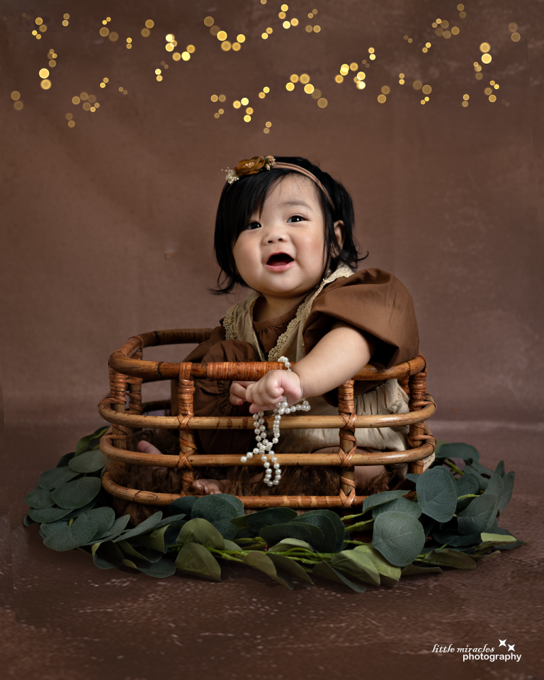 Happy baby sitting in a wicker basket with greenery and gold sparkle background, sitter session photography in Doha