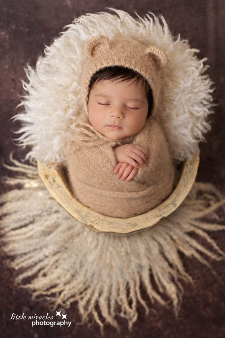 Sleeping newborn in a knitted bear hat curled in a wooden bowl on fluffy fur, newborn photoshoot in Doha