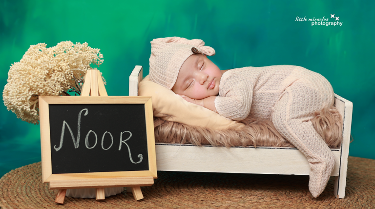 Newborn baby asleep on a small wooden bed in a knitted outfit beside a chalkboard name sign