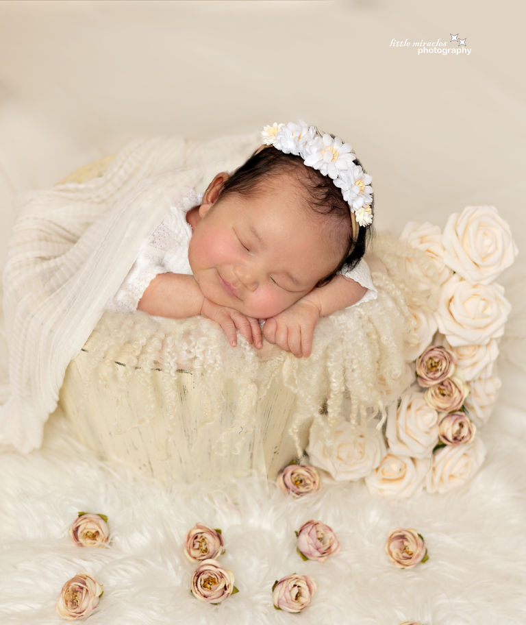 Smiling sleeping newborn girl with a flower headband on a cream bed of roses